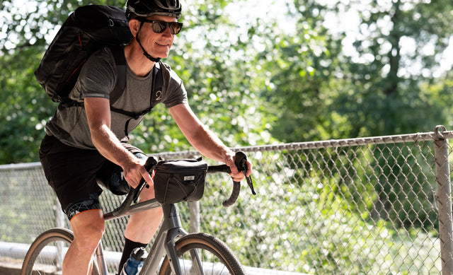 Happy man riding bike wearing the new Two Wheel Gear Spin Backpack and the new Clutch bag attached to handlebars.