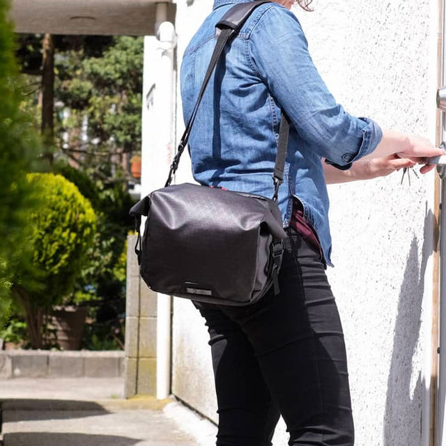 Person wearing a black Two Wheel Gear Alpha handlebar shoulder bag, standing outdoors near a building.