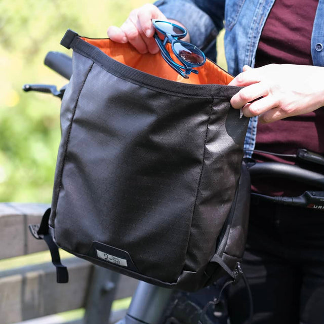 Person holding a black two wheel gear alpha handlebar bag with sunglasses on a blurred outdoor background