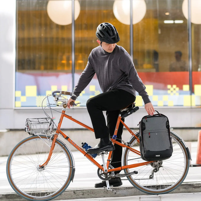 Person on an orange bicycle with a grey two wheel gear pannier backpack plus 2.1, helmet, and water bottle in an urban setting.