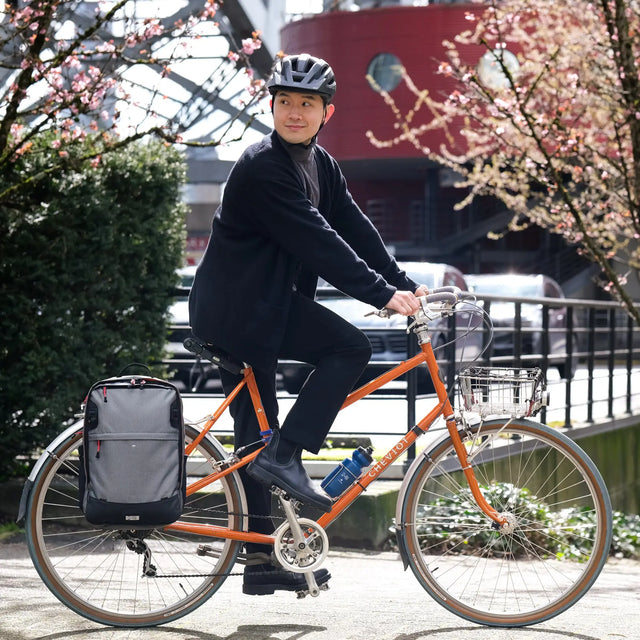 Man riding an orange bicycle with a gray two wheel gear pannier backpack and helmet, cherry blossoms in the background.