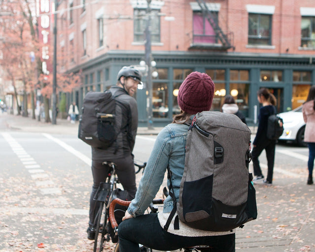 Man and woman riding bikes, both wearing Two Wheel Gear Commute Backpack.